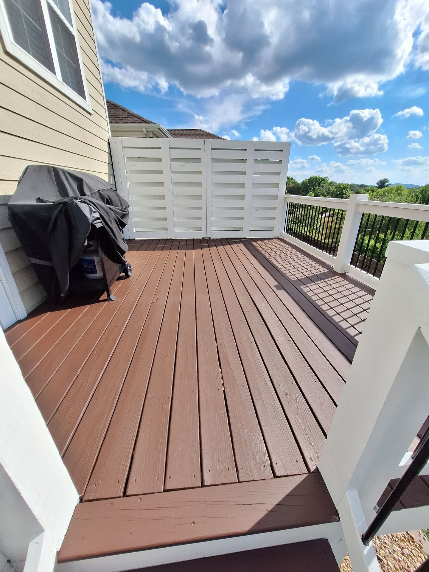 Wooden deck with barbecue grill, white fence, and view of cloudy sky and greenery.